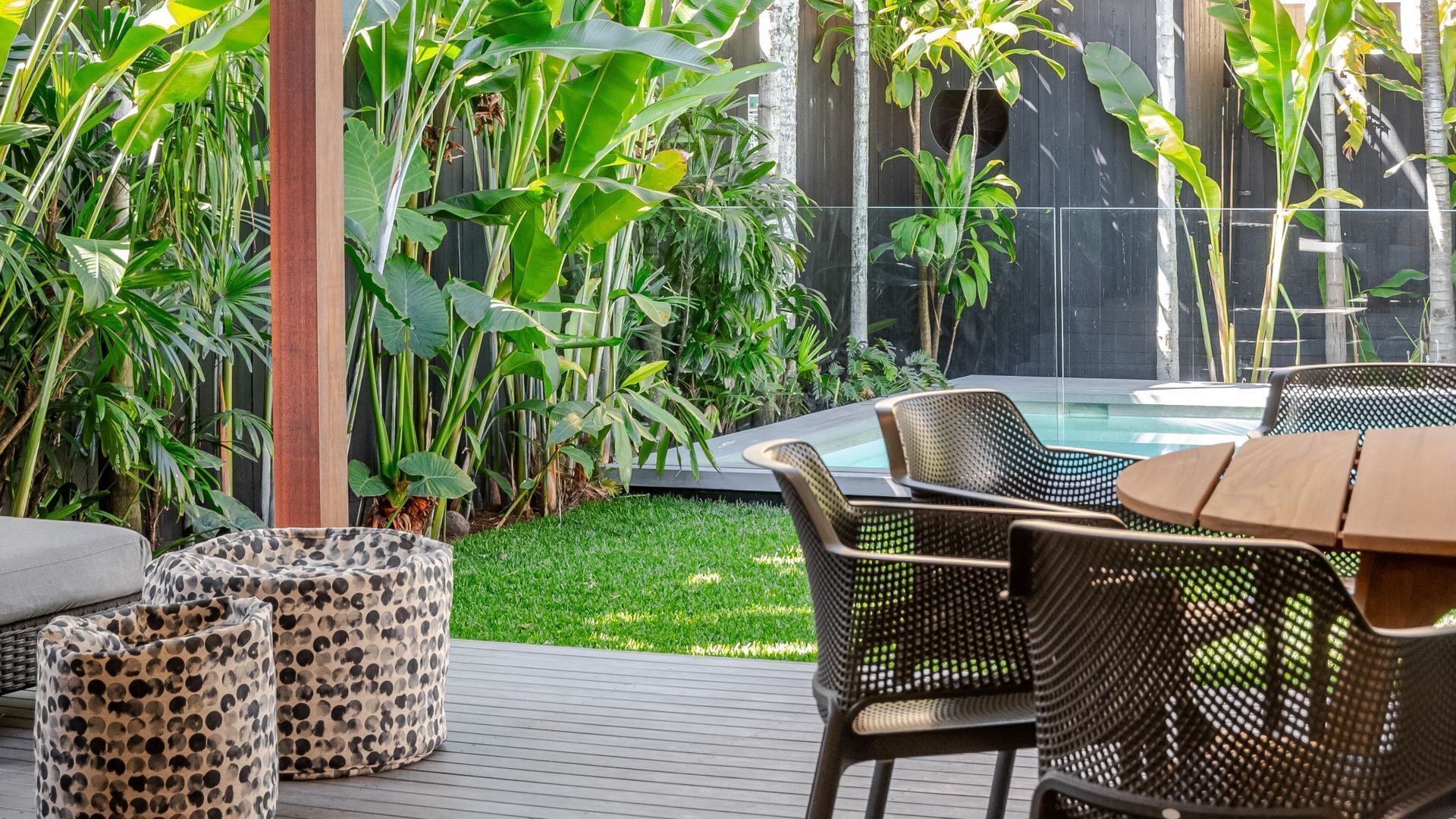 Une terrasse extérieure avec une piscine, entourée d’une végétation tropicale luxuriante et aménagée avec des fauteuils en osier et une table en bois.