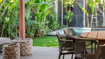 Une terrasse extérieure avec une piscine, entourée d’une végétation tropicale luxuriante et aménagée avec des fauteuils en osier et une table en bois.