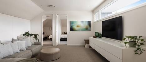 Una sala de estar moderna con un sofá gris, un reposapiés y un televisor de pantalla plana grande sobre un mueble blanco. Hay un cuadro en la pared y una planta sobre el mueble.