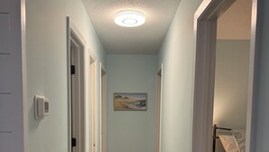 A narrow hallway with wooden flooring, light blue walls, and a ceiling light. There is a door on the left and a bed with patterned bedding on the right.