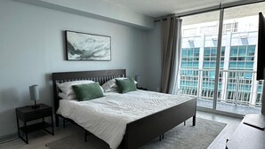 Un dormitorio moderno con cama, mesita de noche, lámpara, ventana grande y vistas de edificios.