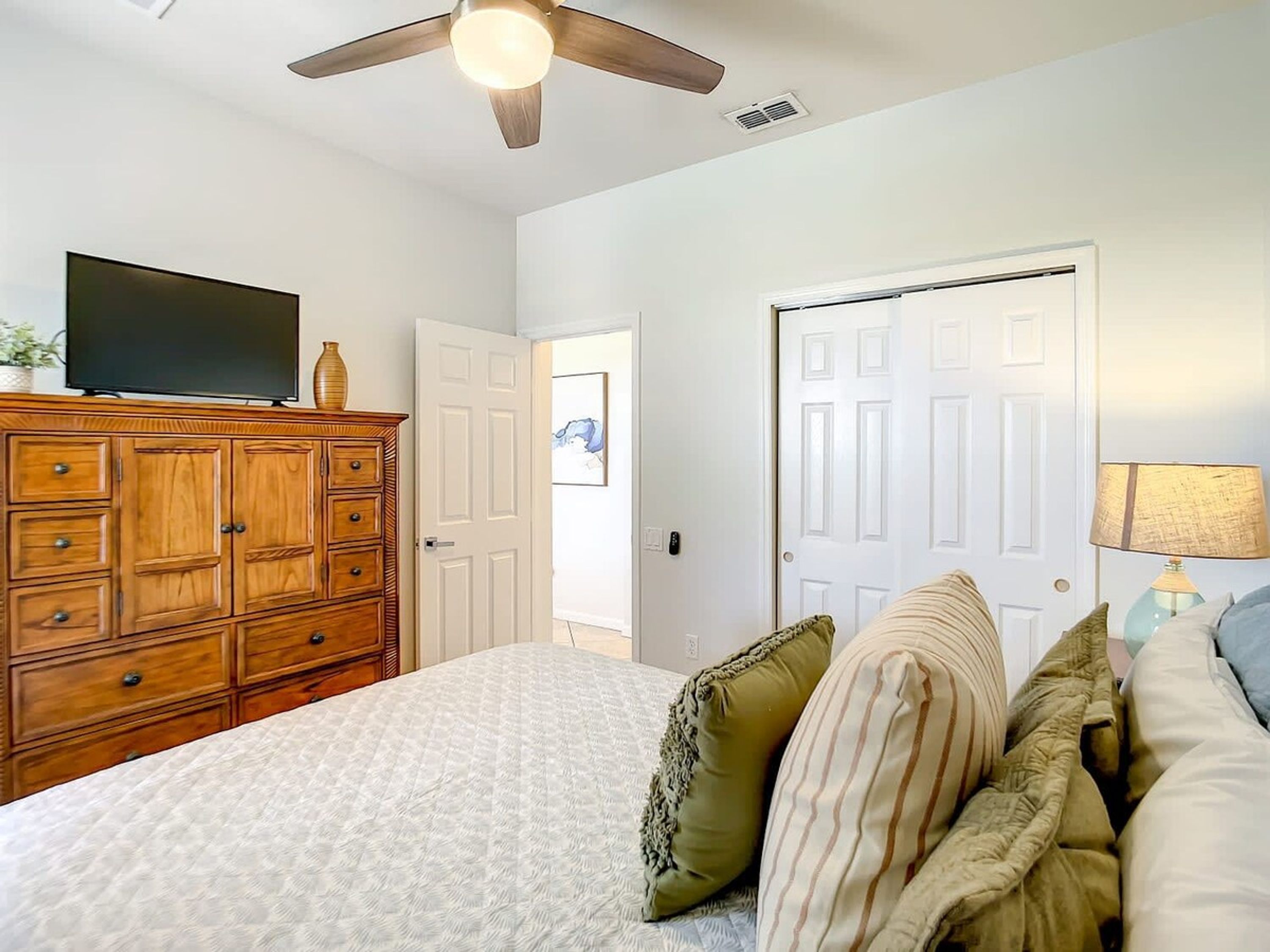 A bedroom with a bed, a wooden dresser, a ceiling fan, a television on the dresser, and a door with a picture on the wall.