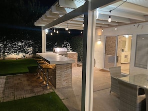 An outdoor kitchen area with a grill, bar stools, and a glass enclosure leading to a patio with a dining table and chairs. - Stunning 5 Star Home Retreat Private Pool Spa BBQ 4 BR (La Quinta)