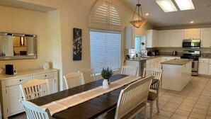 A dining area with a long table, white chairs, a chandelier, and a kitchen with white cabinets and appliances. - Stunning 5 Star Home Retreat Private Pool Spa BBQ 4 BR (La Quinta)