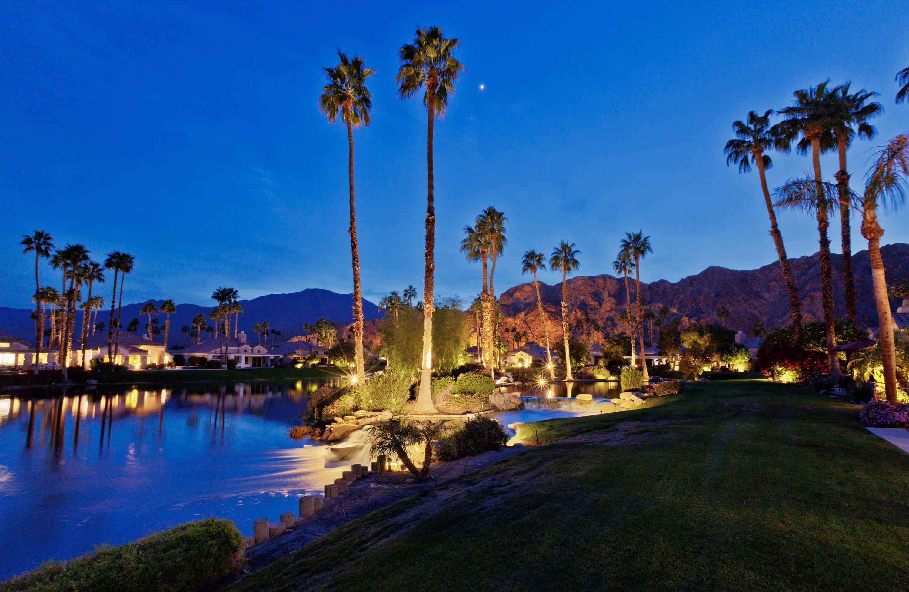 A nighttime view of a golf course with palm trees, a lake, and mountains in the background.