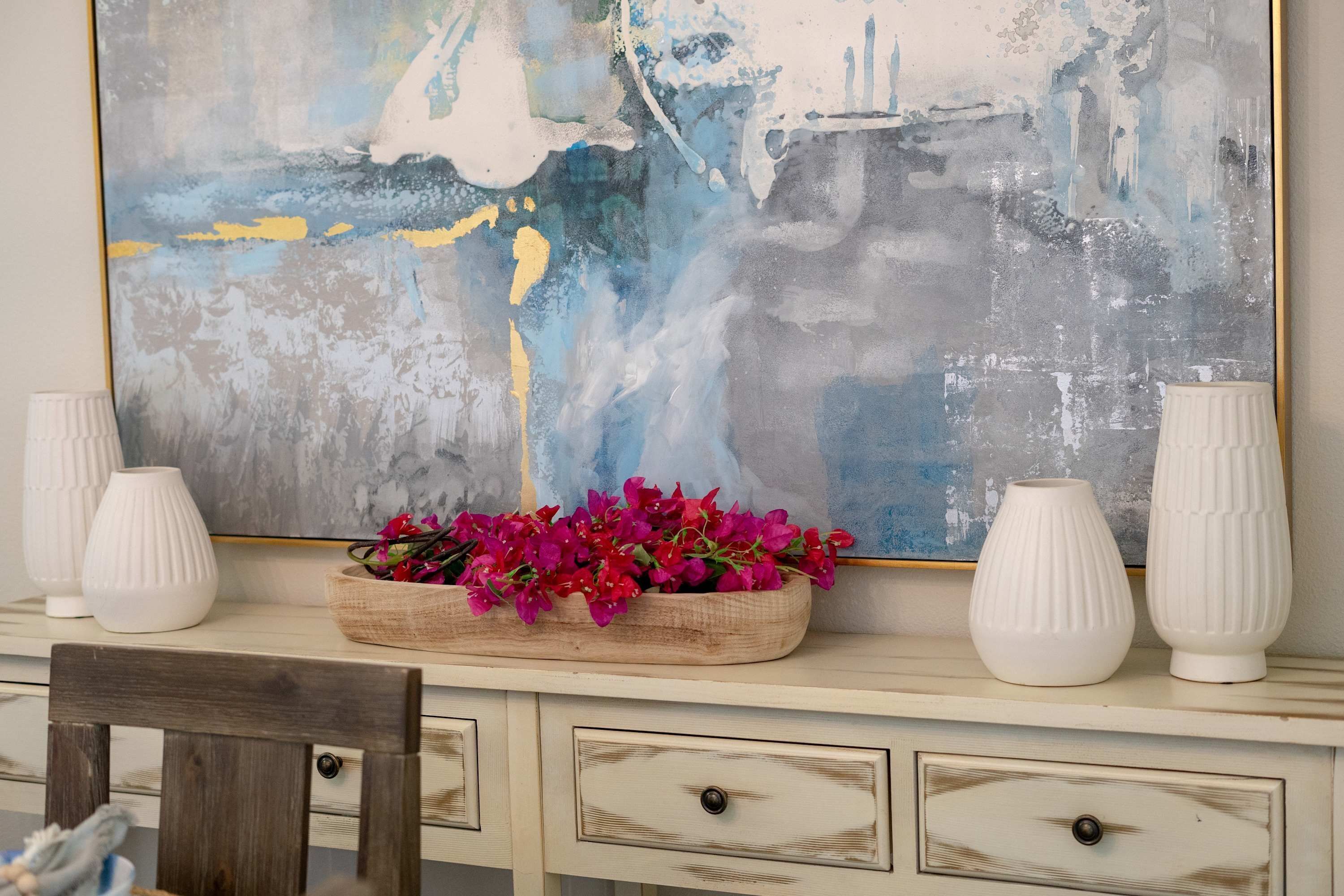 Un bureau surmonté d’un grand tableau abstrait, un plateau en bois avec des fleurs roses et deux vases blancs.