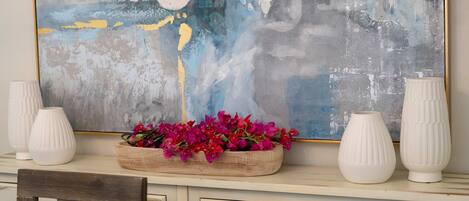 Un bureau surmonté d’un grand tableau abstrait, un plateau en bois avec des fleurs roses et deux vases blancs.