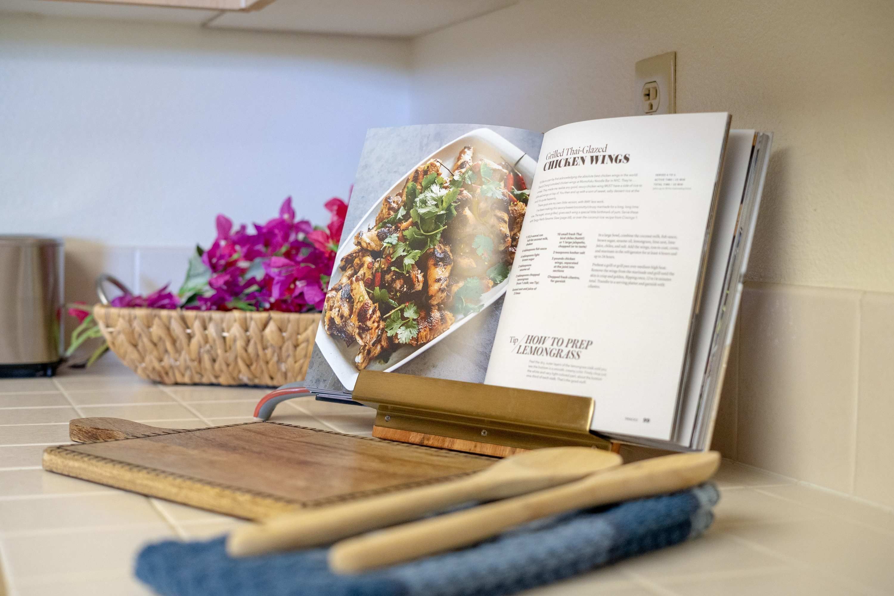 A cookbook open on a kitchen counter, with a wooden cutting board and a pair of knives beside it.