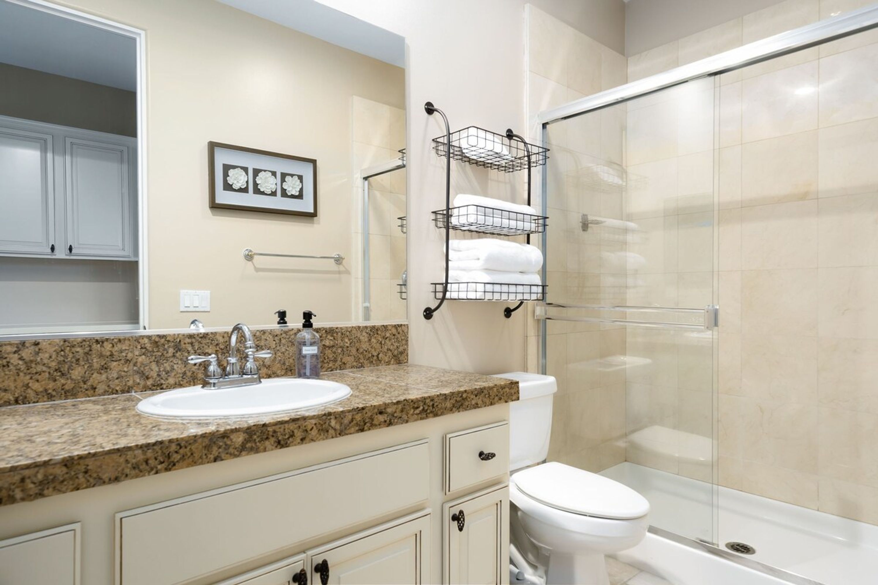 A bathroom with a granite countertop sink, a white toilet, a glass shower enclosure, and a towel rack with folded towels.