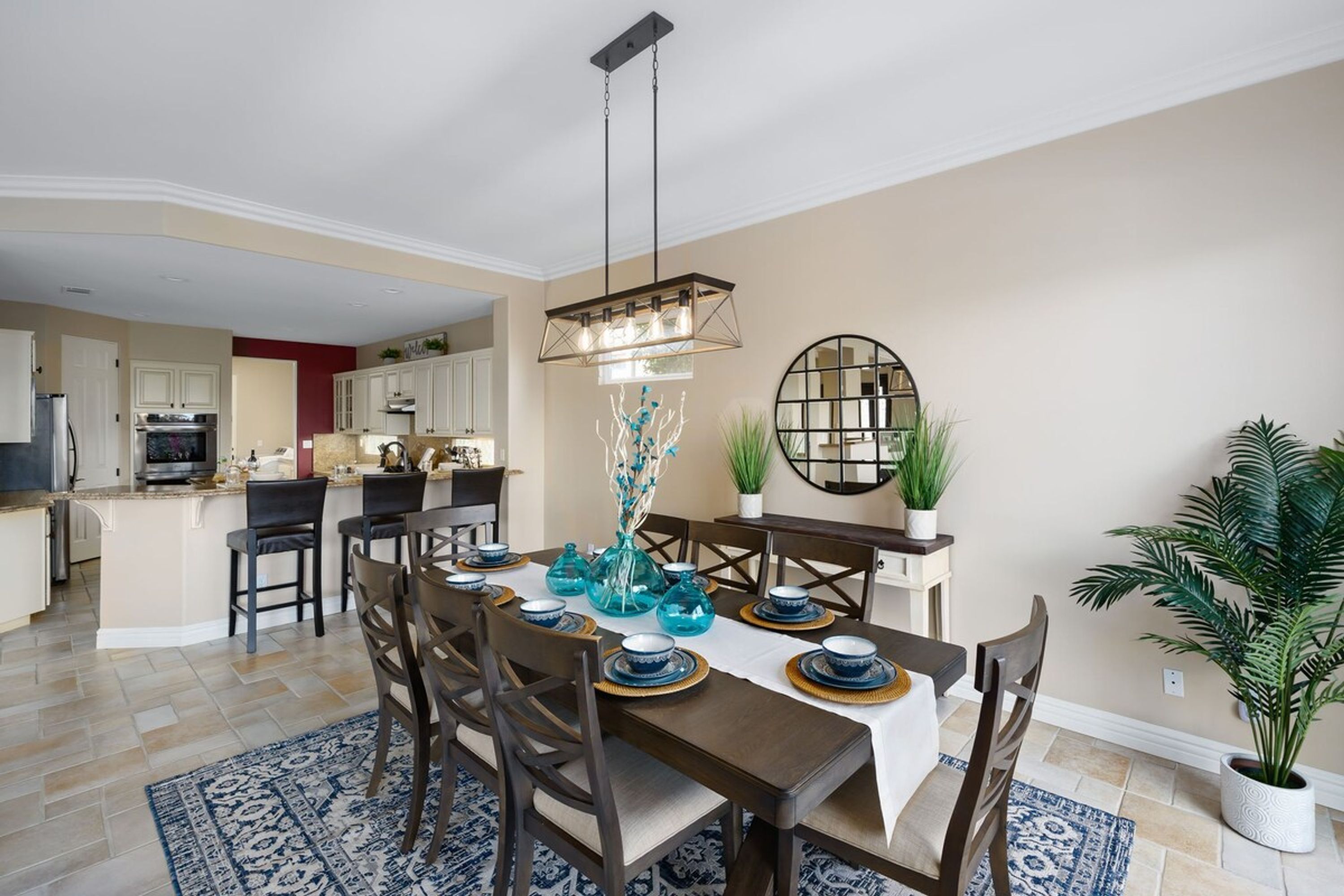 A dining area with a long table set for six, surrounded by chairs. There is a chandelier above the table, and a potted plant is visible to the side.