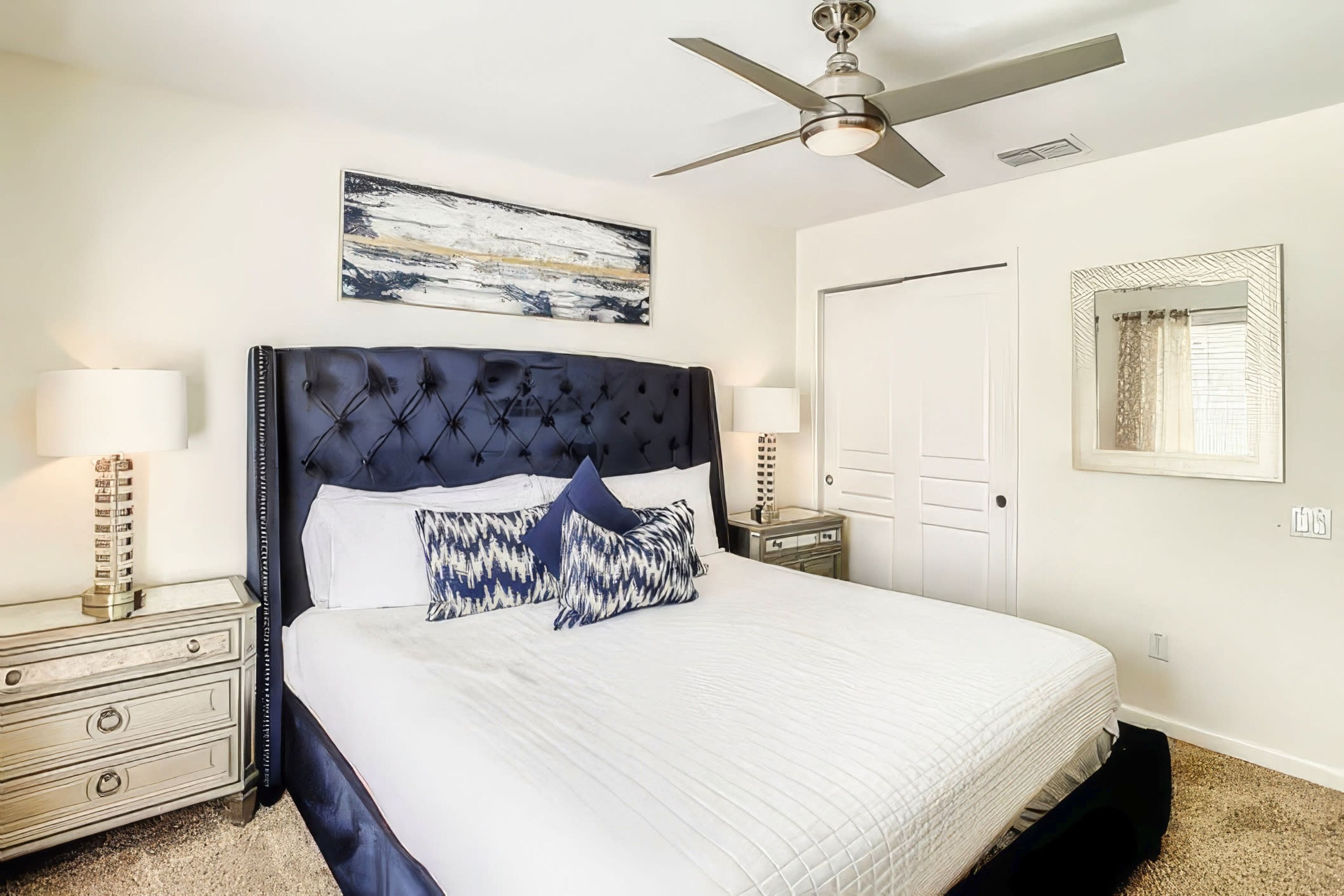 A bedroom with a large bed, a ceiling fan, a nightstand with a lamp, a dresser, and a door.