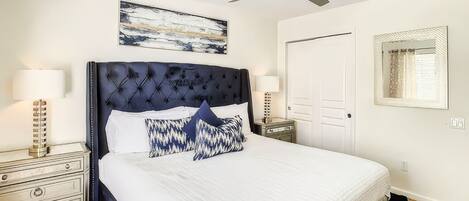A bedroom with a large bed, a ceiling fan, a nightstand with a lamp, a dresser, and a door.