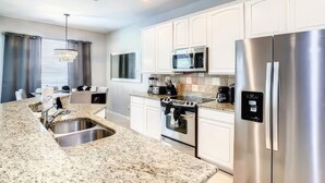 A modern kitchen with white cabinets, stainless steel appliances, granite countertops, and a dining area with a chandelier. - Ultimate Adventure Villa Top Rated 5 Star Disney More (Kissimmee)