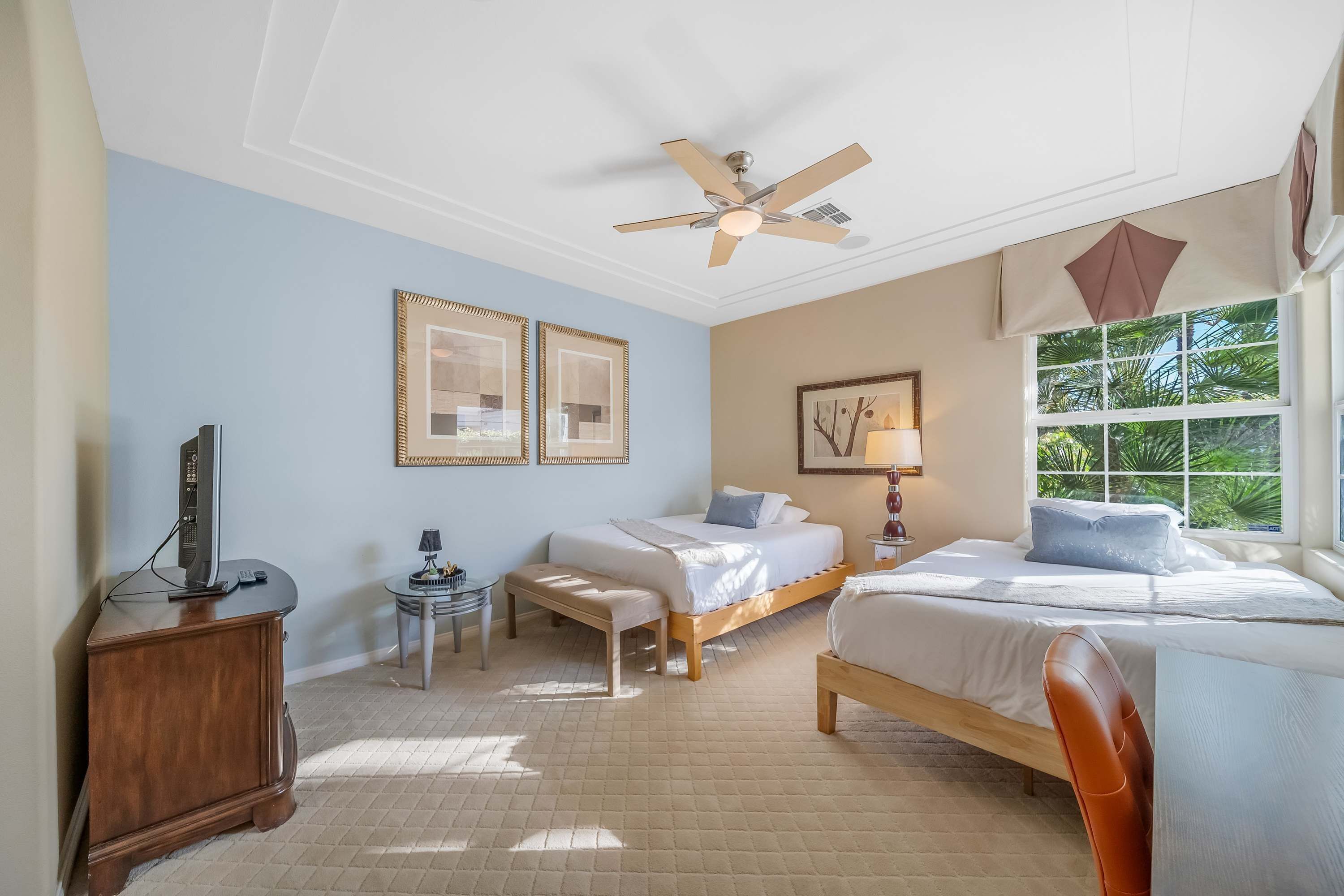 A hotel room with a bed, a desk, a chair, a ceiling fan, and a window with a view of greenery.