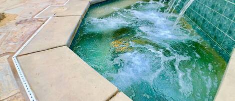 A rectangular hot tub with a waterfall feature, surrounded by a tiled patio.