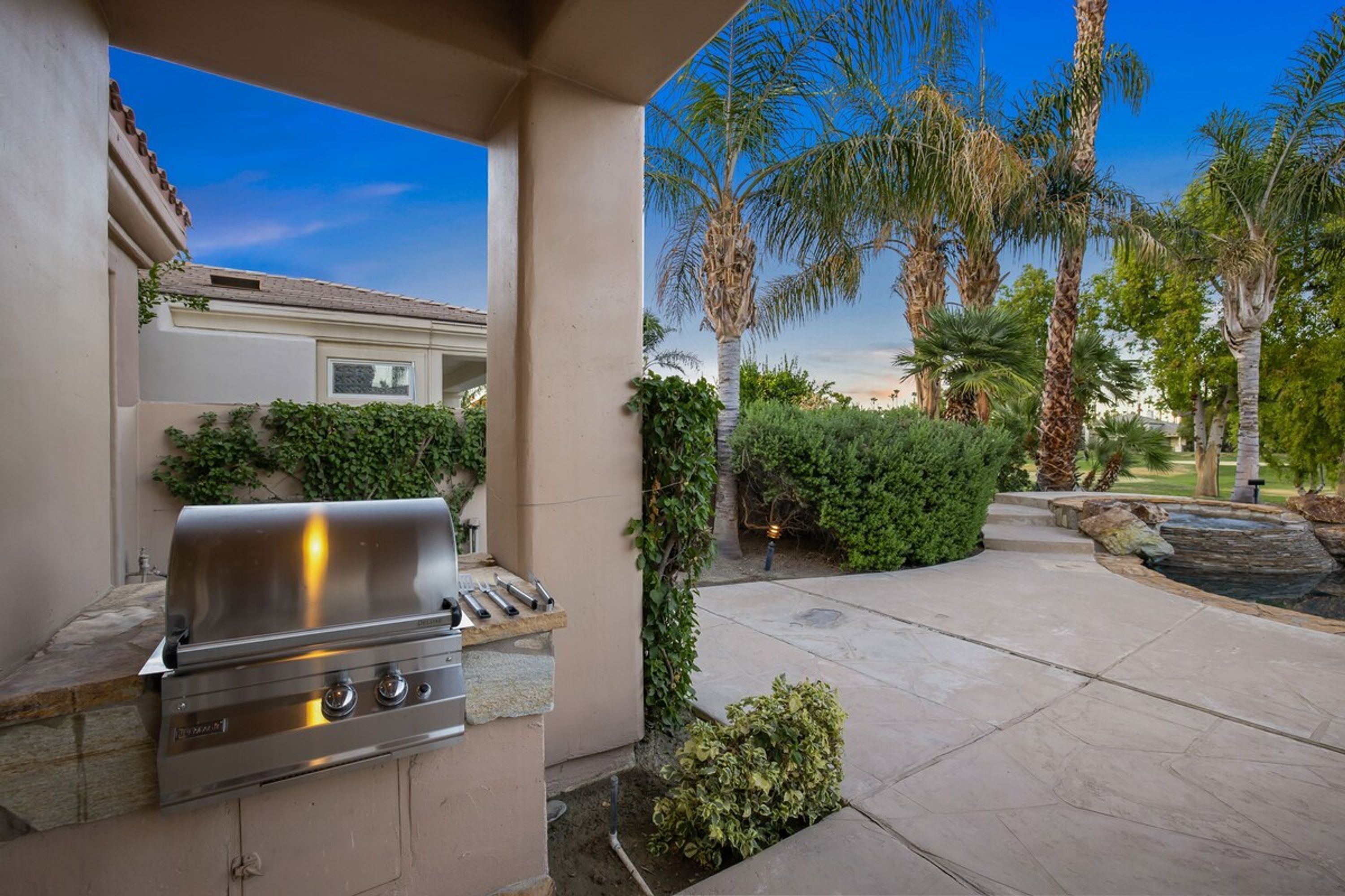 An outdoor patio with a built-in grill, surrounded by palm trees and a stone pathway.