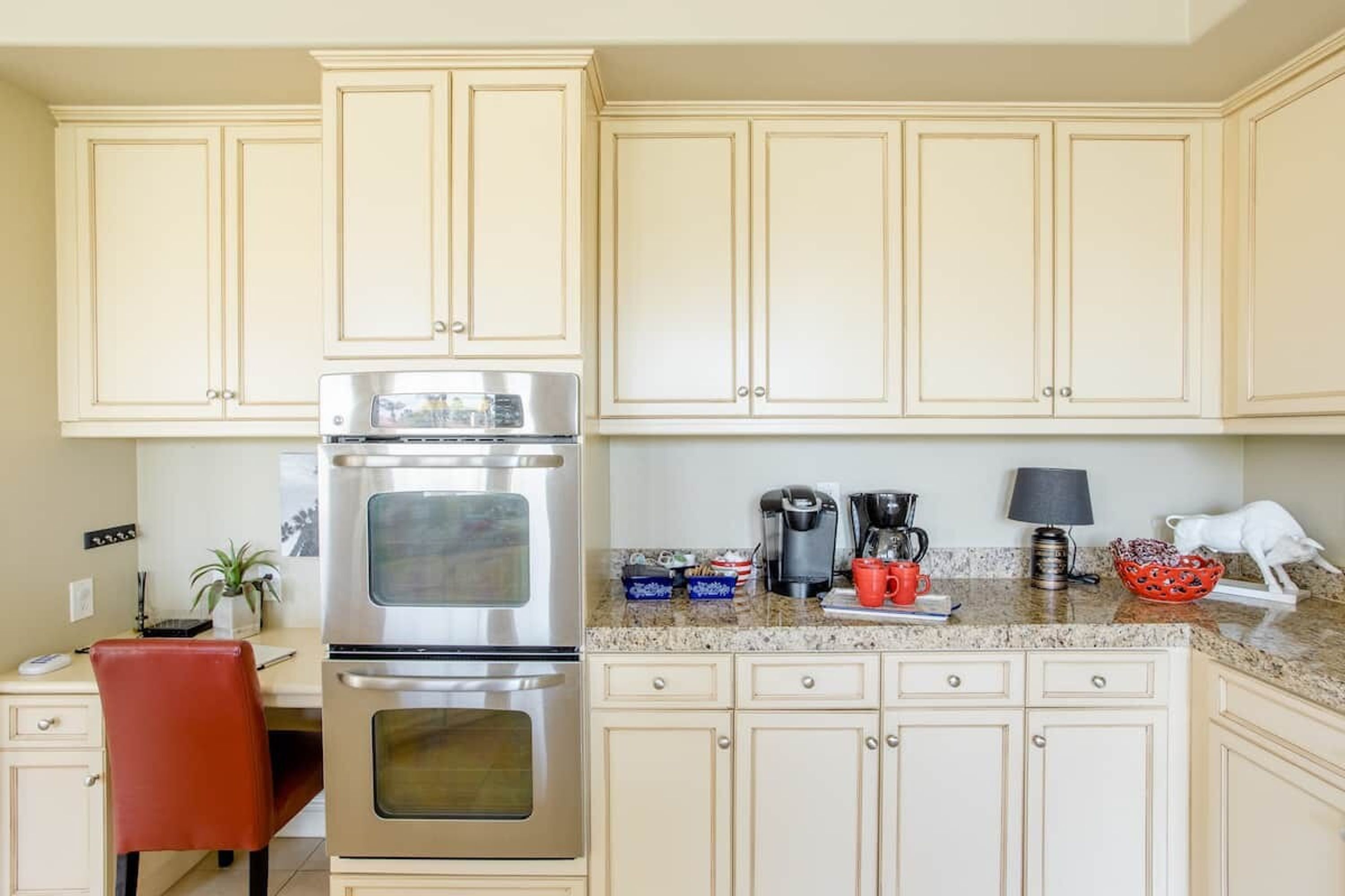 A kitchen with cream-colored cabinets, a stainless steel oven, a coffee maker, and a red mug on the countertop.