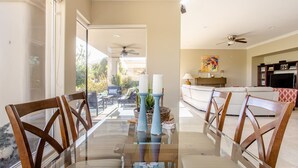 A dining area with a glass table and wooden chairs, a kitchen in the background, and a view of the outdoors. - Casa Santiago Private Pool Firepit Golf View (La Quinta)