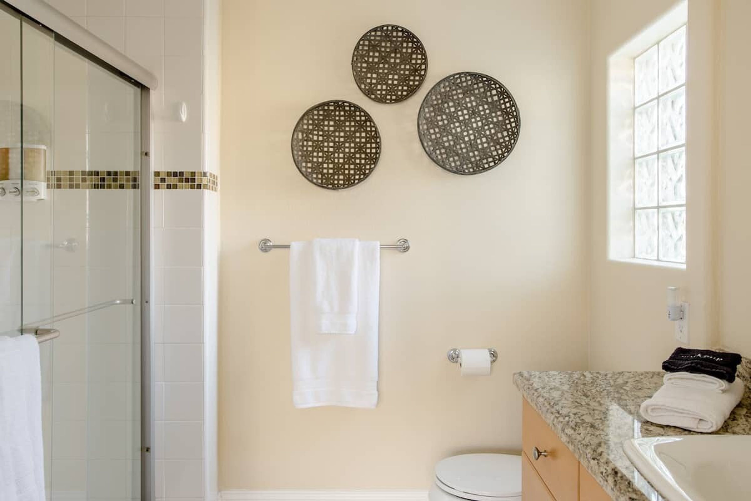 Un baño con cabina de ducha de vidrio, inodoro y lavamanos con encimera de granito. Se observan tres adornos circulares en la pared sobre el inodoro.