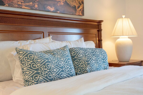 A neatly made bed with white linens and two patterned pillows, a wooden headboard, and a bedside lamp on a nightstand. - Charming Condo With Golf Course View (Palm Desert)