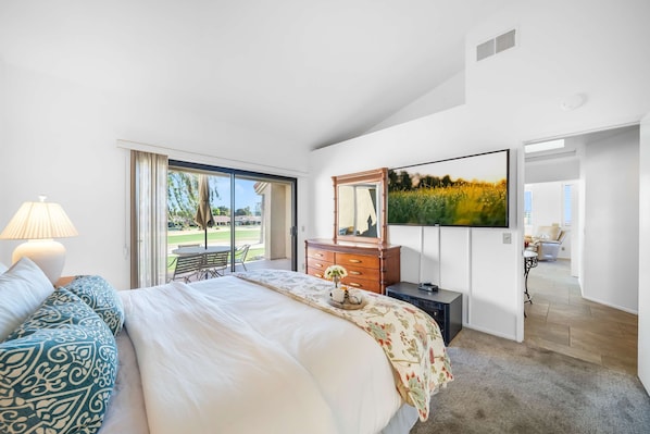A modern bedroom with a large bed, a dresser, a TV, and a view of the outdoors. - Charming Condo With Golf Course View (Palm Desert)