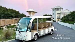 A white beach shuttle with a canopy, labeled "KIVA SHUTTLE," parked on a paved path with a beach house in the background.