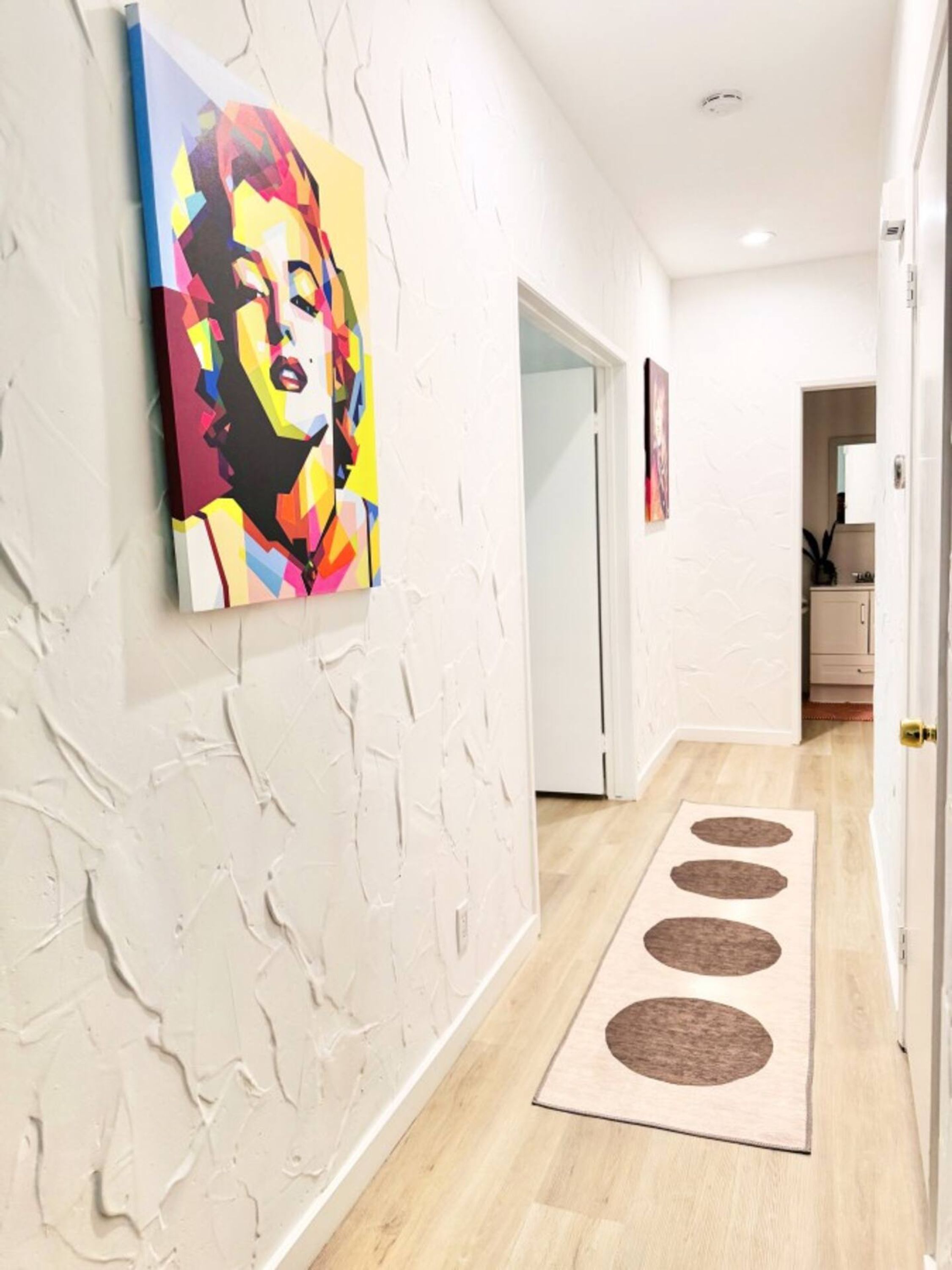 Een gang met een witte, gestructureerde muur, een kleurrijk pop-art schilderij en een houten vloer met een bruin vloerkleed.