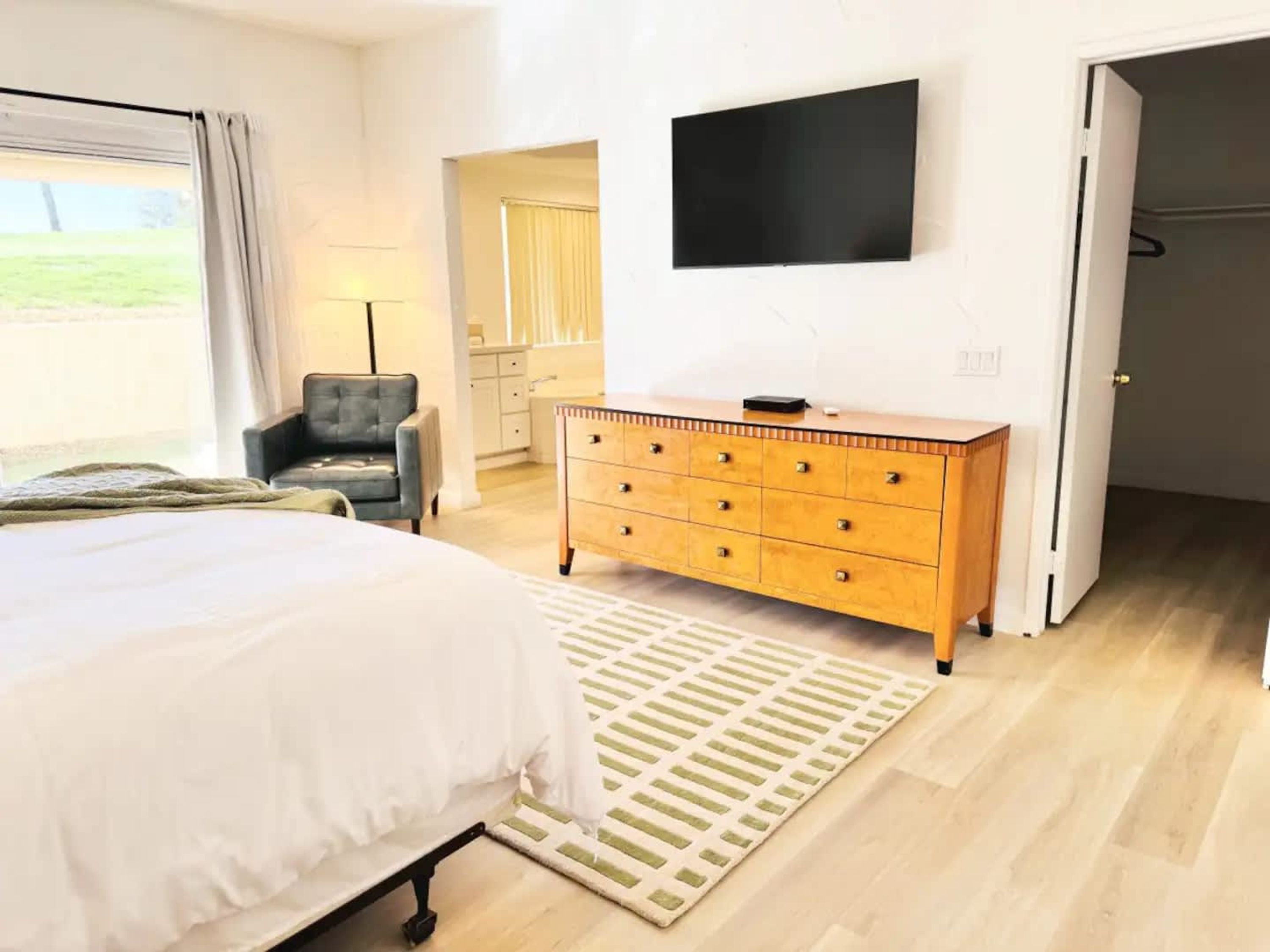 Un dormitori amb un llit, una petita taula de nit, un televisor fixat a la paret i una porta que condueix a una altra habitació.