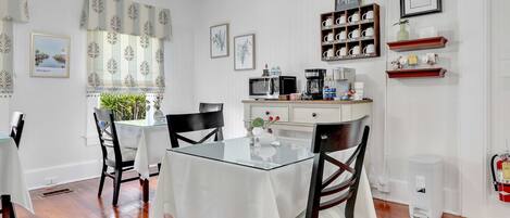 A dining area with a glass table, chairs, a coffee machine, and a shelf with mugs and cups.