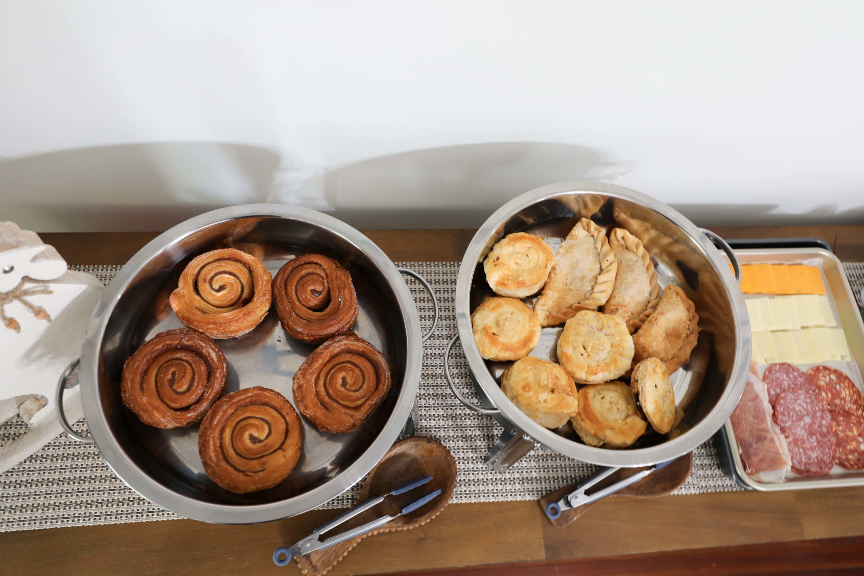 A table with two metal pans, one containing cinnamon rolls and the other containing pastries and meat.