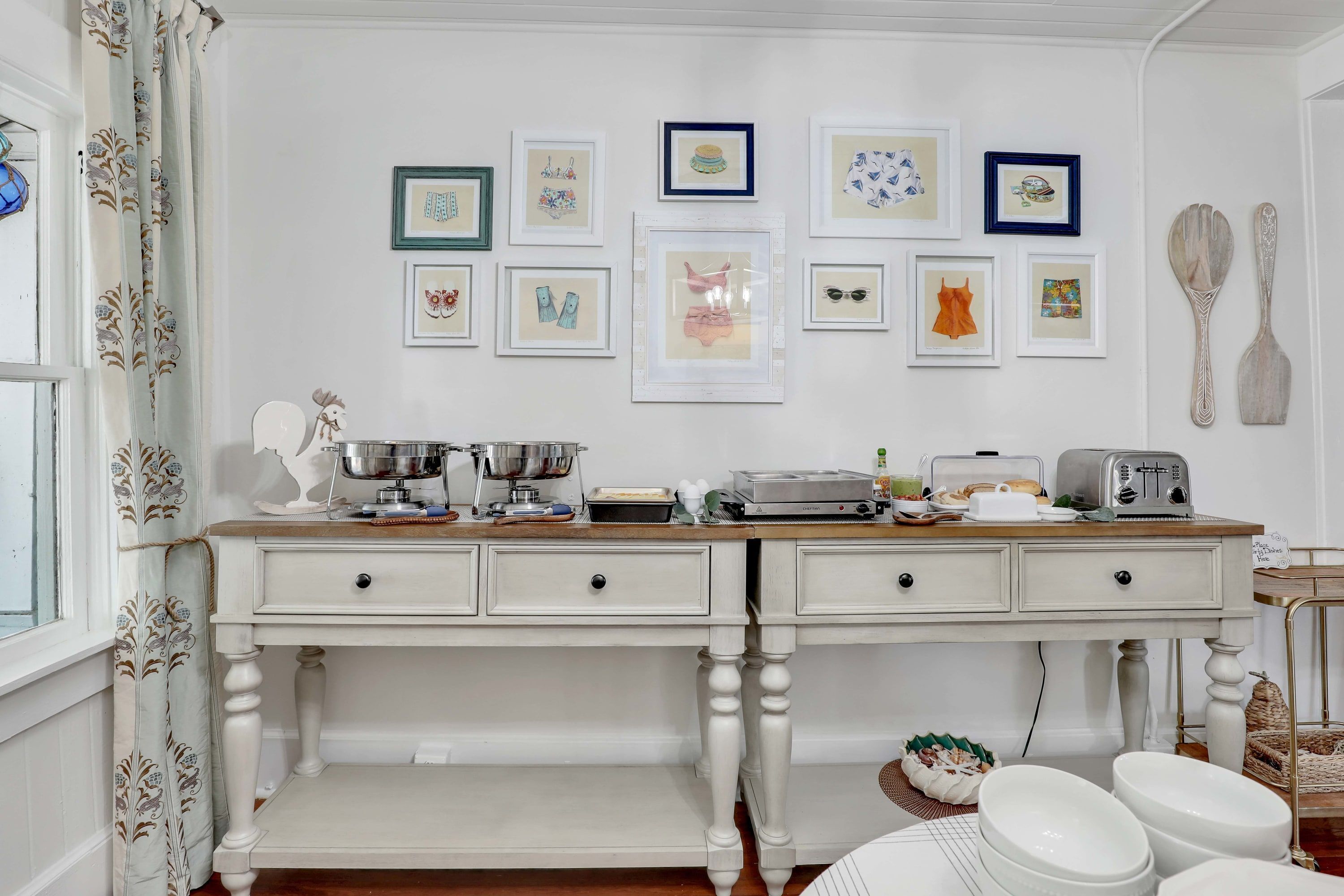 A kitchen with a white double sink, a stove, and a countertop with various appliances and utensils. There are framed pictures on the wall above the counter.