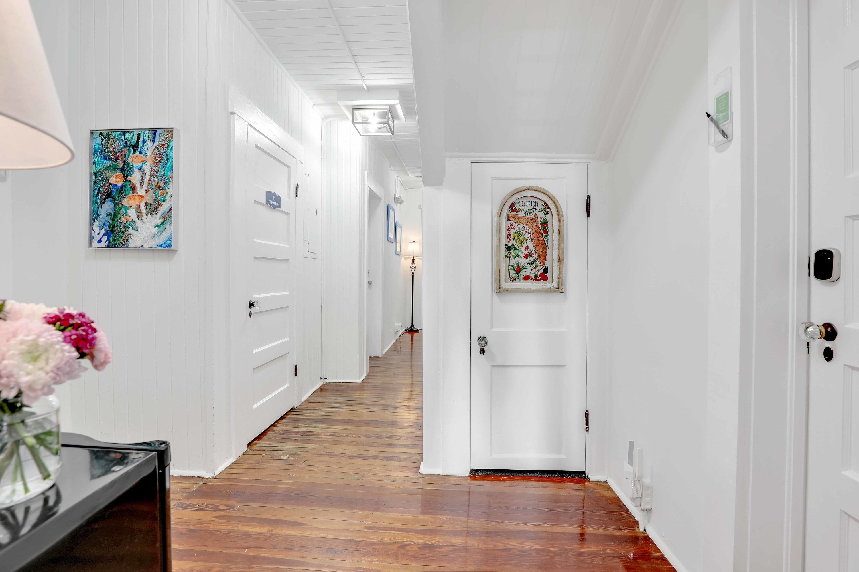 Un pasillo con paredes blancas, piso de madera y una puerta con arco decorativo. Se ve un cuadro en la pared, y un florero con flores sobre una mesa.