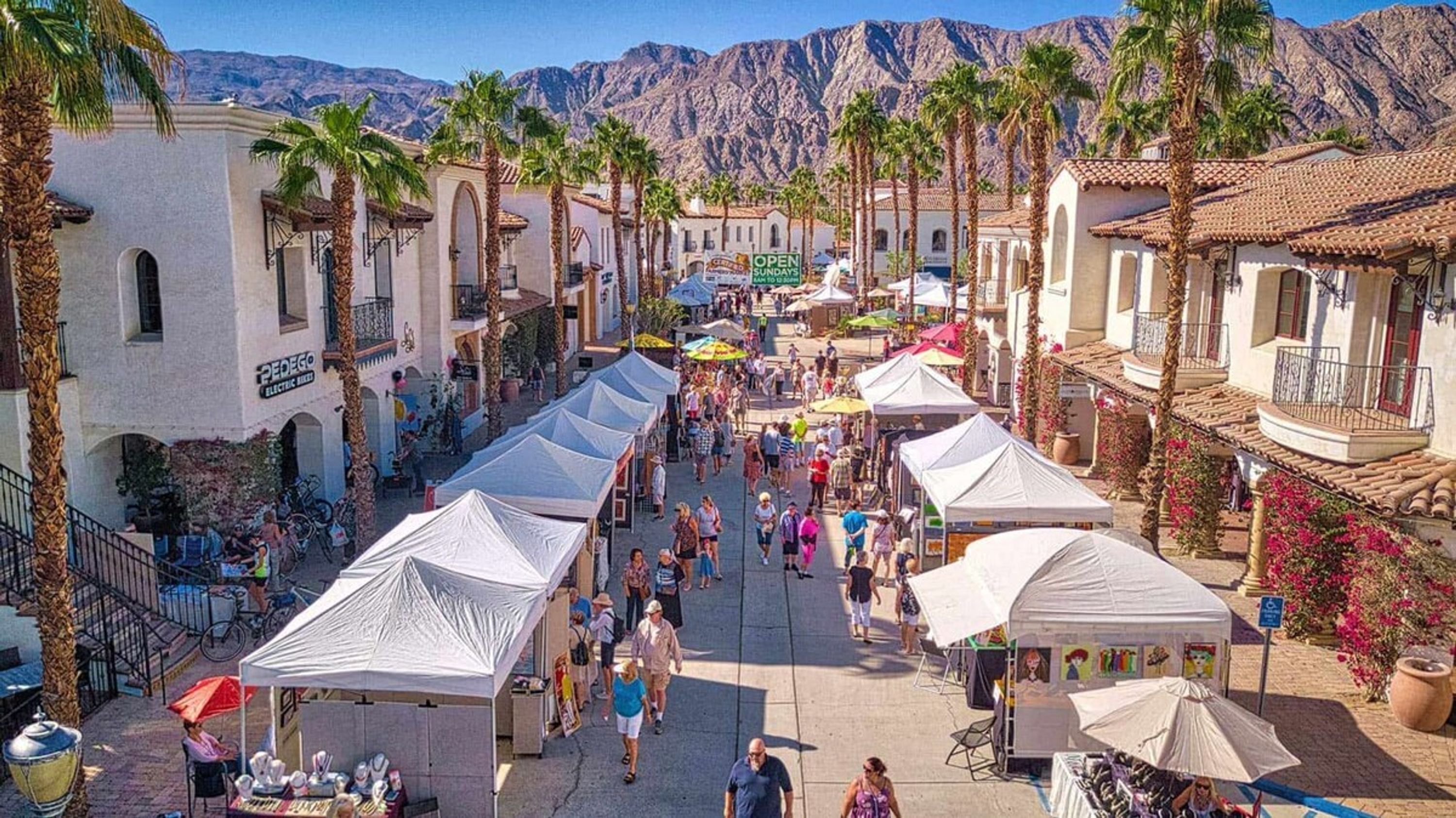 A bustling pedestrian street lined with palm trees and market stalls, with mountains in the background.