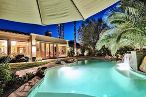 A swimming pool with a waterfall feature, surrounded by palm trees and a covered patio area.