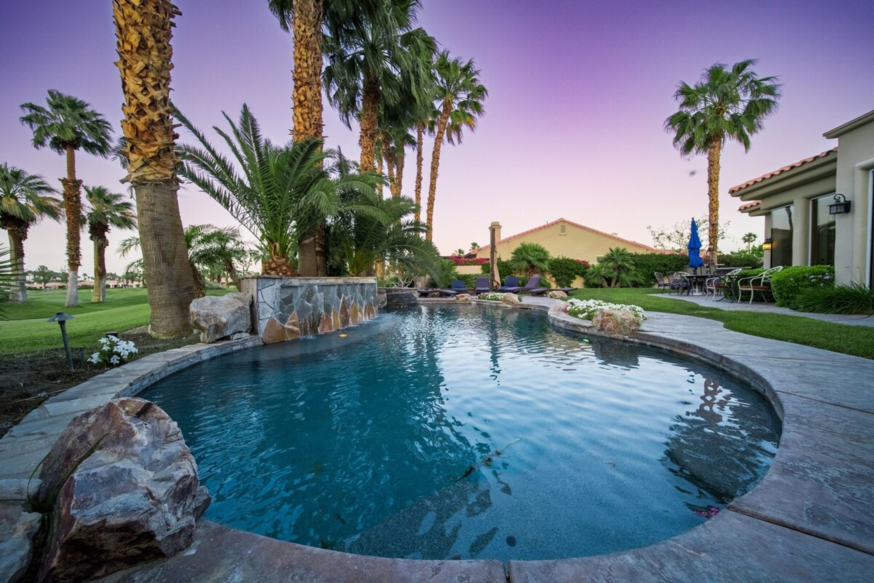 A circular pool with a stone border, surrounded by palm trees and a well-maintained lawn.