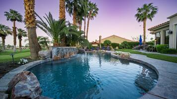 A circular pool with a stone border, surrounded by palm trees and a well-maintained lawn.