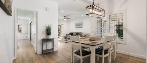 A modern dining area with a wooden table and chairs, a hanging light fixture, and a view of a living room in the background.