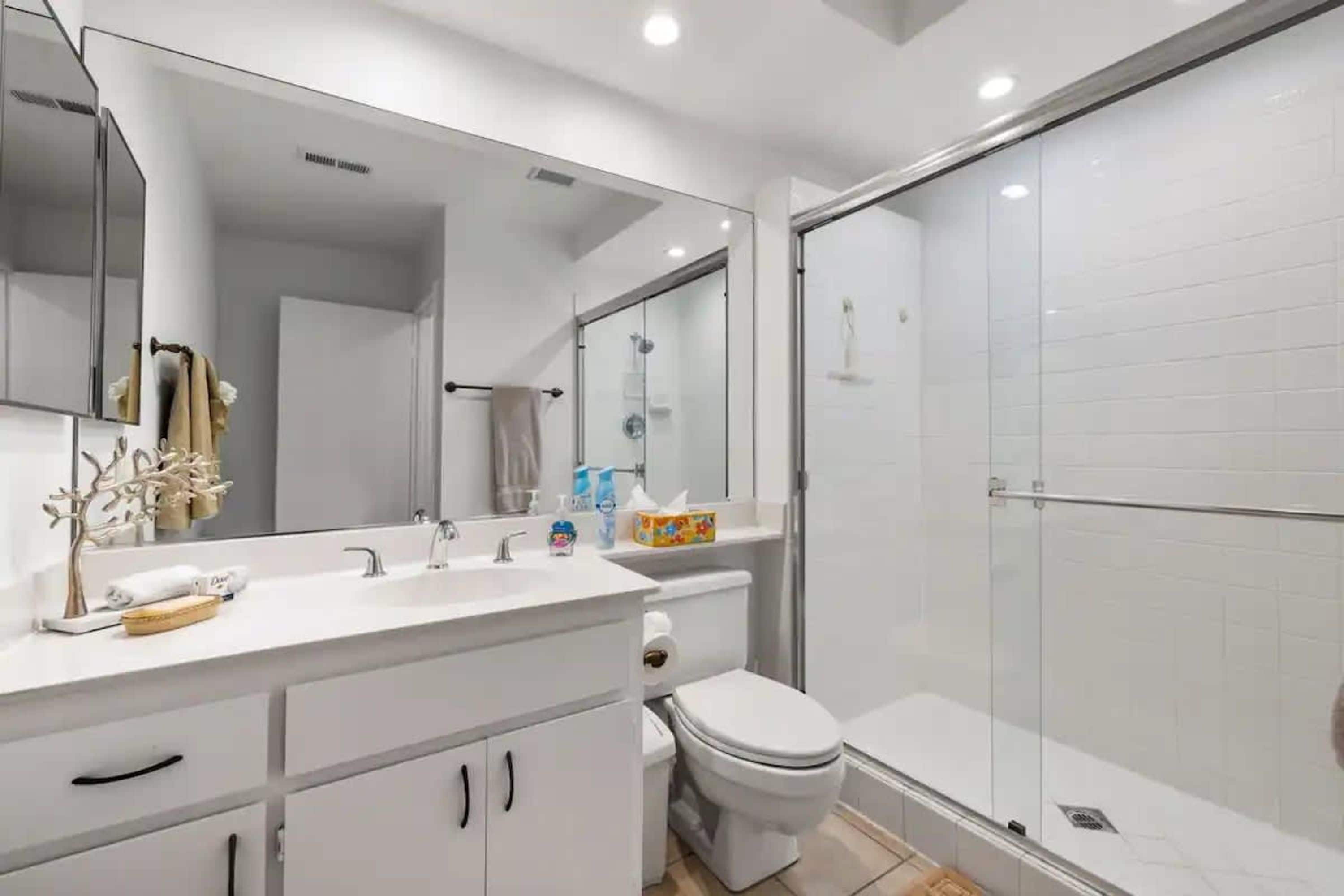 A modern bathroom with a large mirror, white cabinets, a toilet, and a glass-enclosed shower.