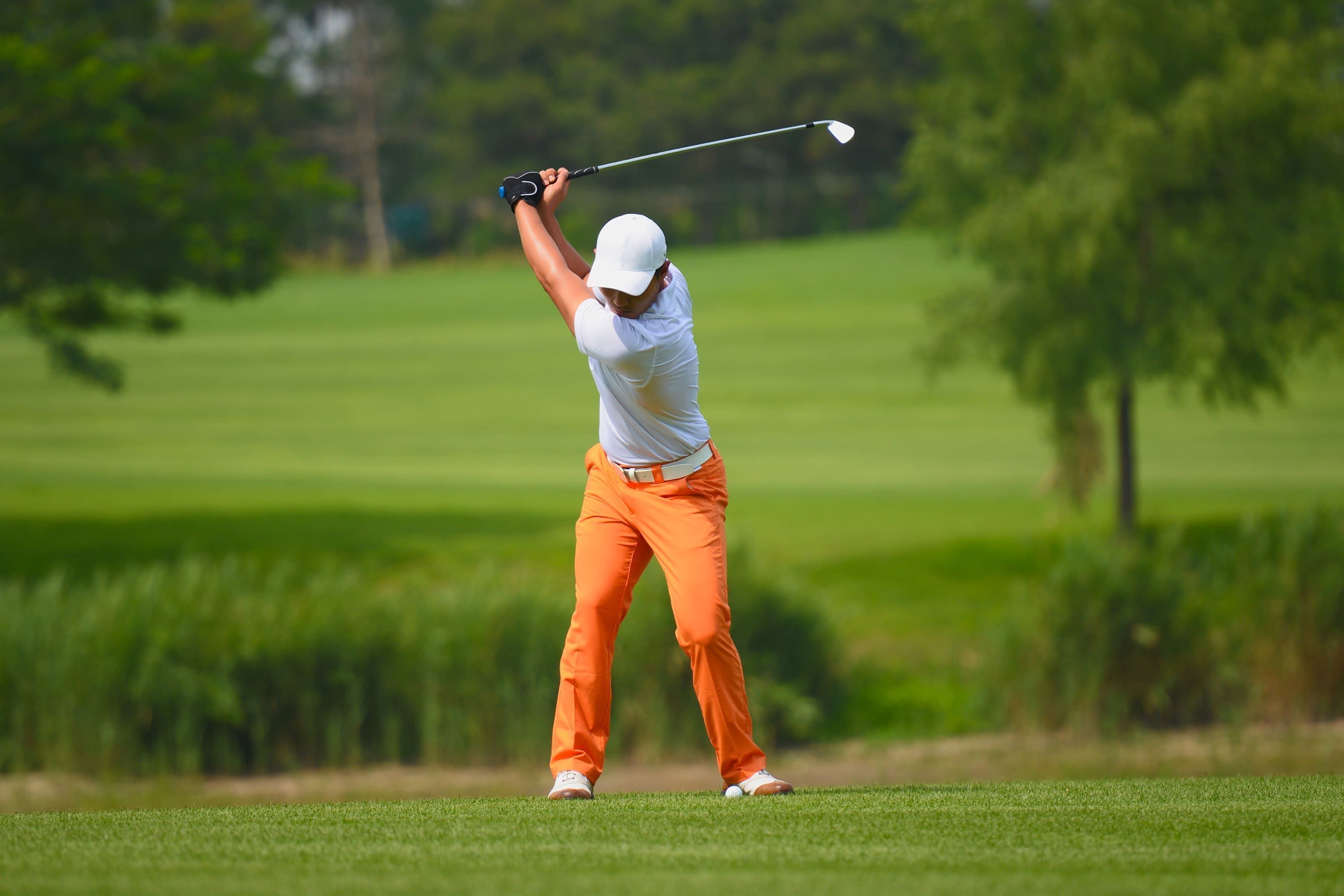 Un golfista en pleno balanceo en un campo de golf, con cielo despejado y árboles al fondo.
