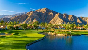 Un campo de golf con un obstáculo acuático, casas y montañas al fondo.