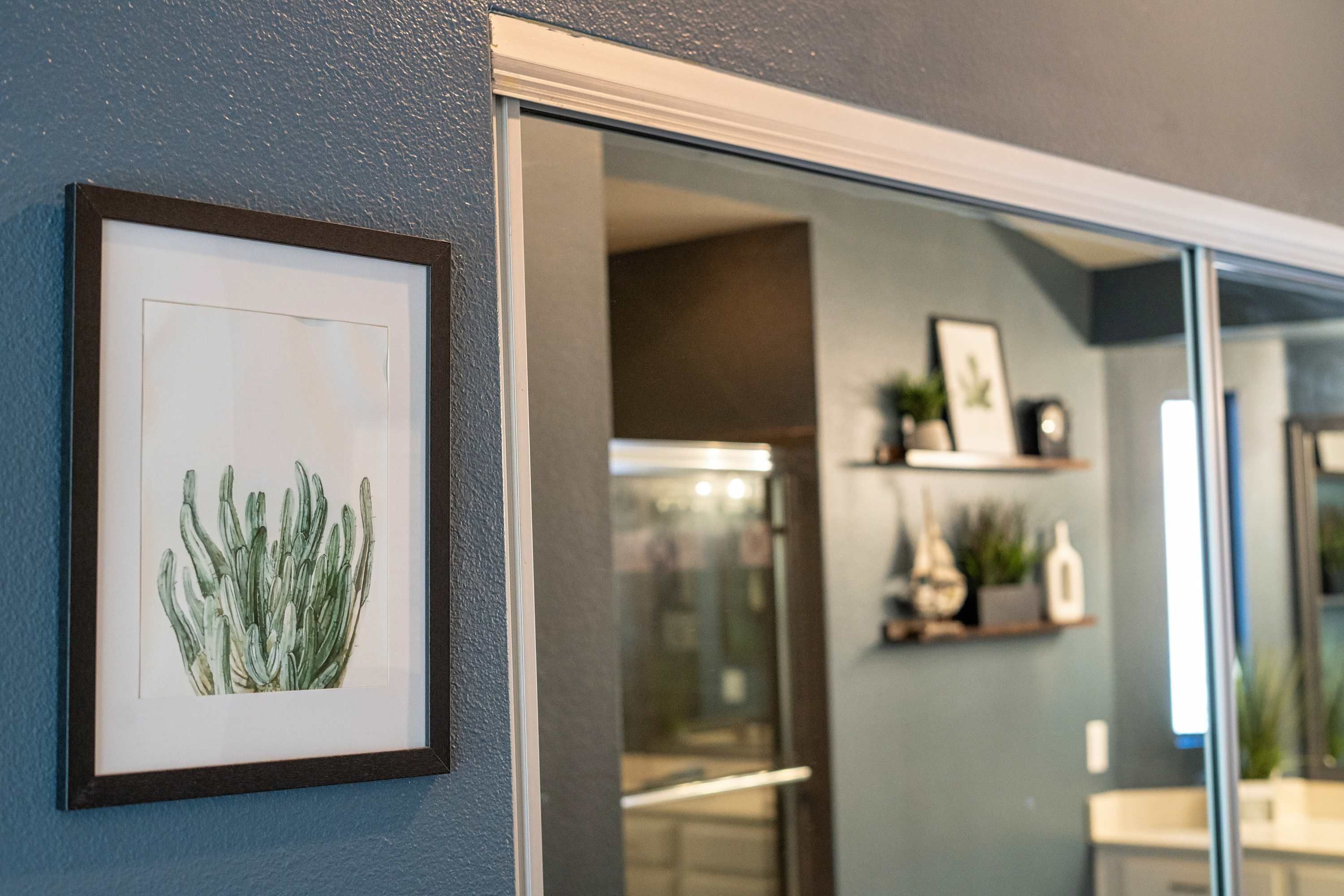 A framed botanical illustration on a wall, a doorway leading to a room with a mirror, and a shelf with decorative items.