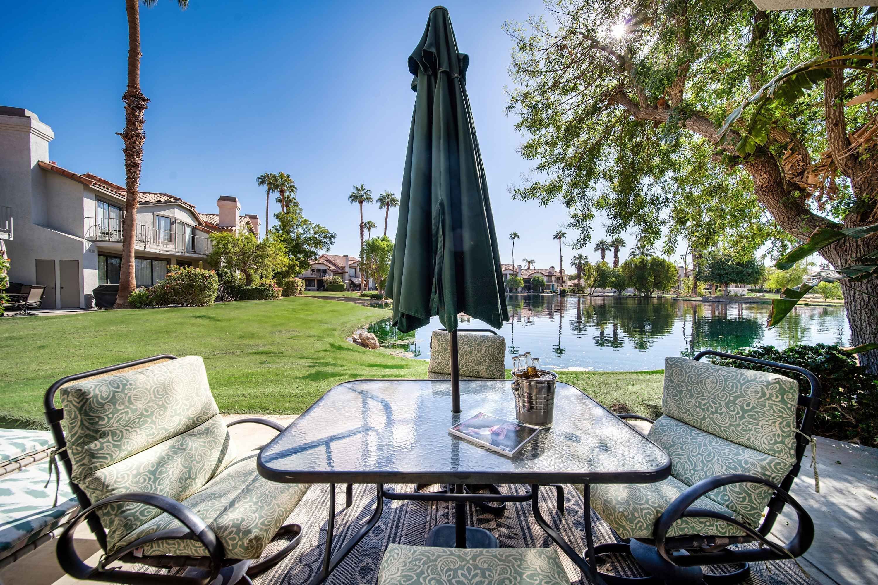 Un patio con una mesa y dos sillas estampadas, un paraguas verde, y una vista de un lago y edificios al fondo.