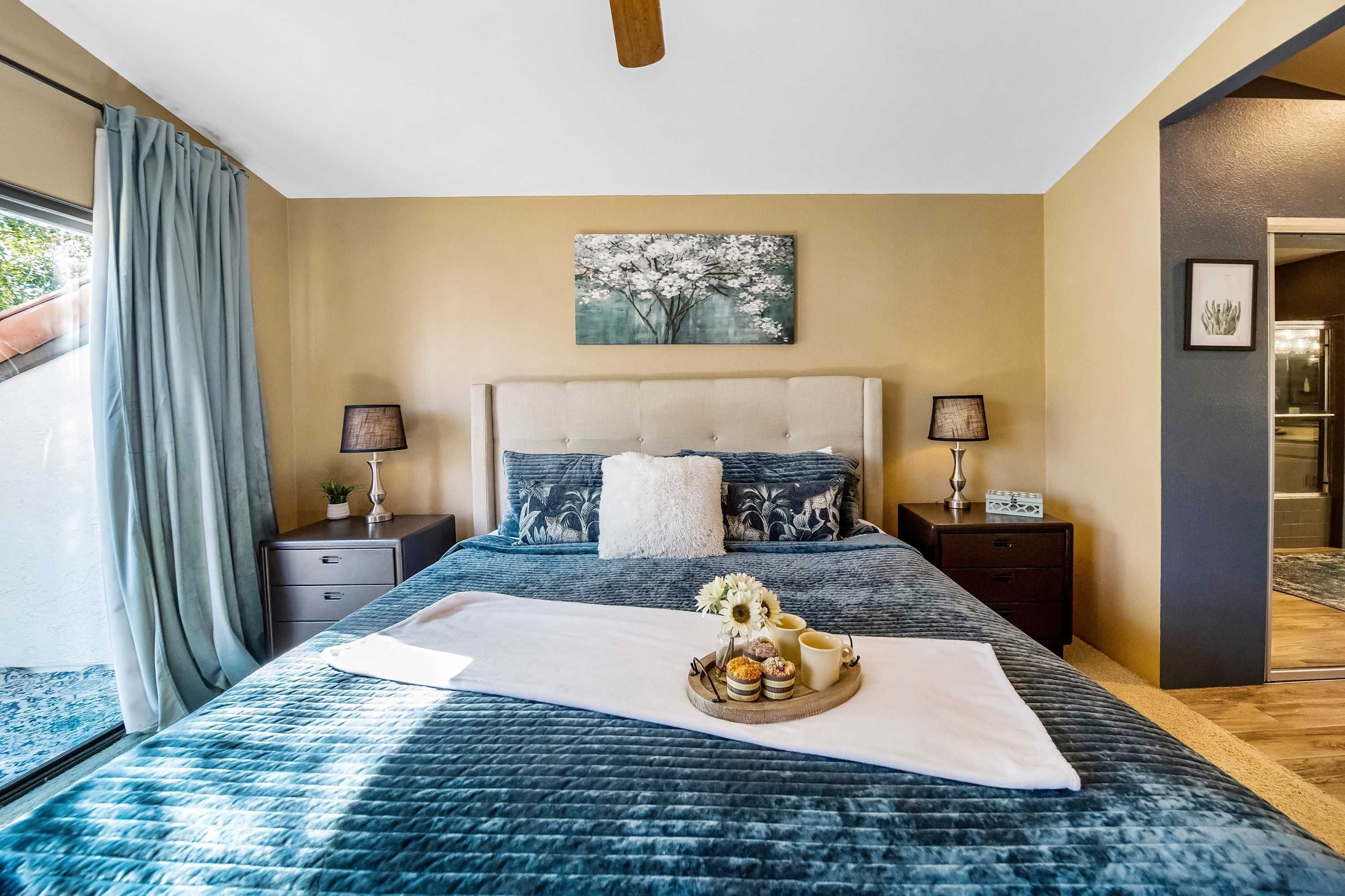 Een slaapkamer met een bed, nachtkastjes, een lamp, een schilderij en een dienblad met spullen.