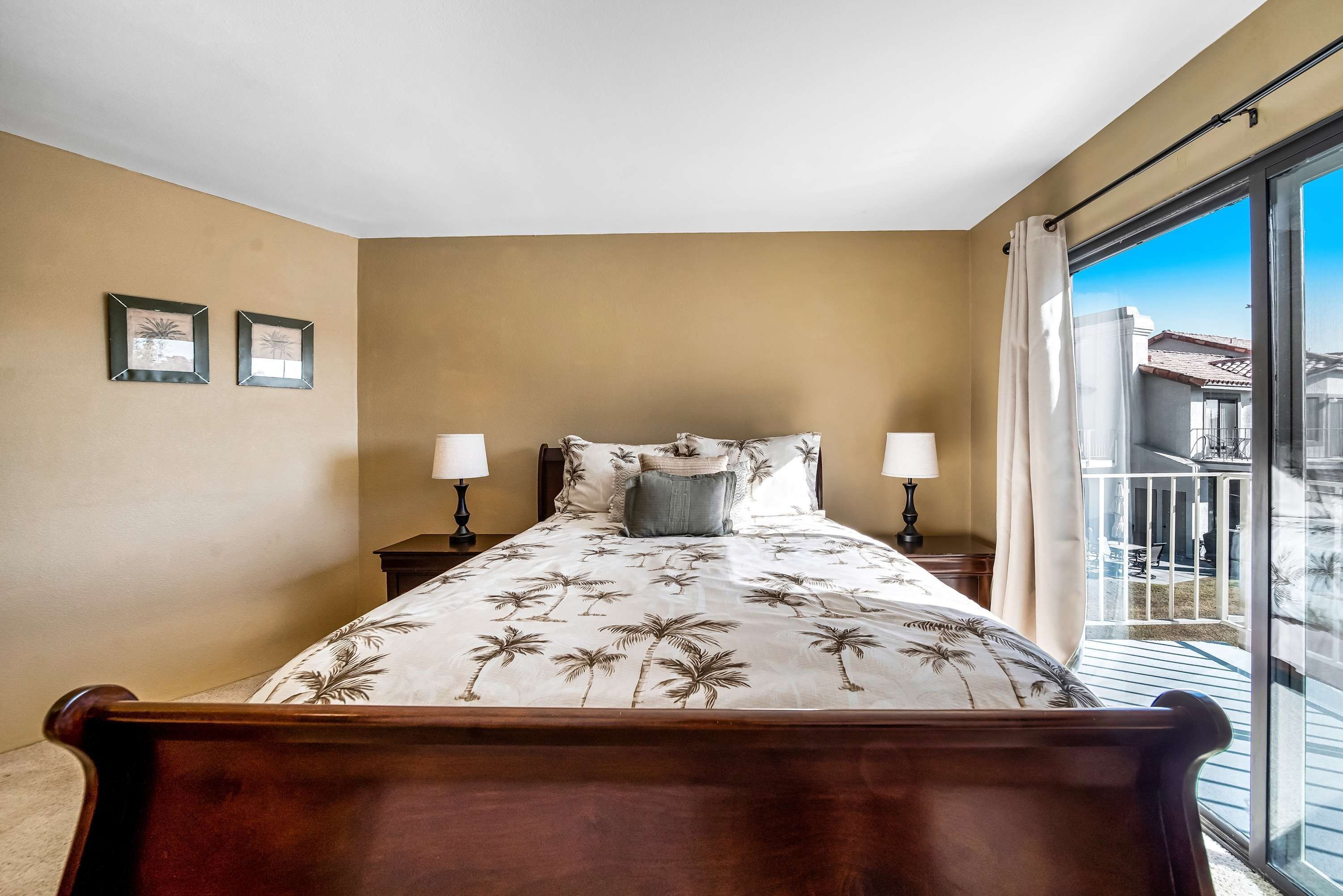 A bedroom with a bed, two nightstands, a balcony with a railing, and a view of buildings.