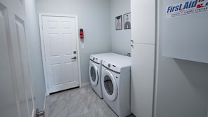 A laundry room with two front-loading washing machines, a white door, and a First Aid sign on the wall. - Palm Luxe Estate Luxury Desert Retreat With Pool (Indio)