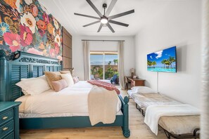 A bedroom with a large bed, a smaller bed, a ceiling fan, a window with curtains, a painting, and a desk with a chair. - Stylish 2BR w Spa Patio (La Quinta)