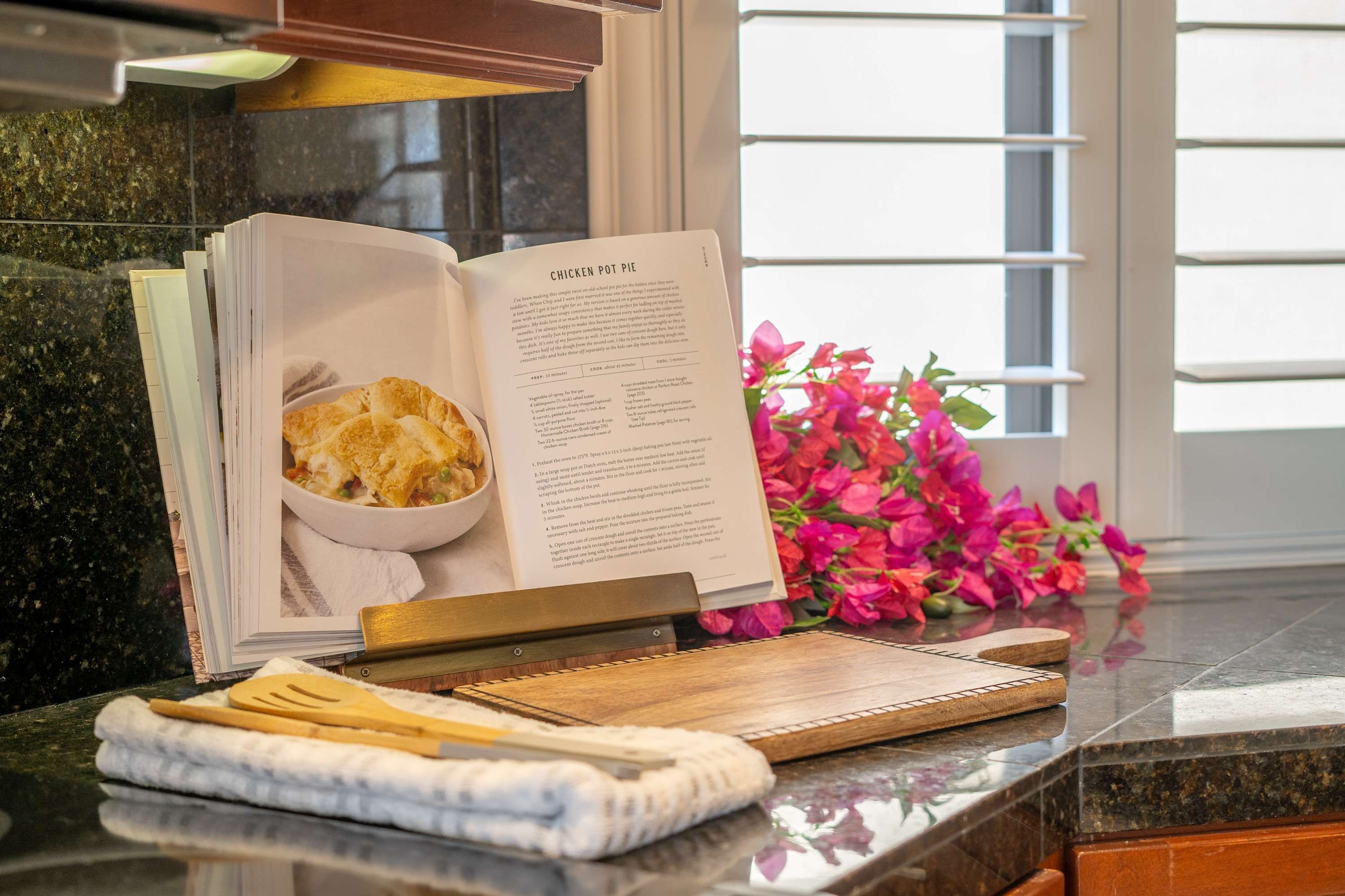 A kitchen counter with a cookbook open to a recipe, a cutting board, and a pair of wooden utensils.