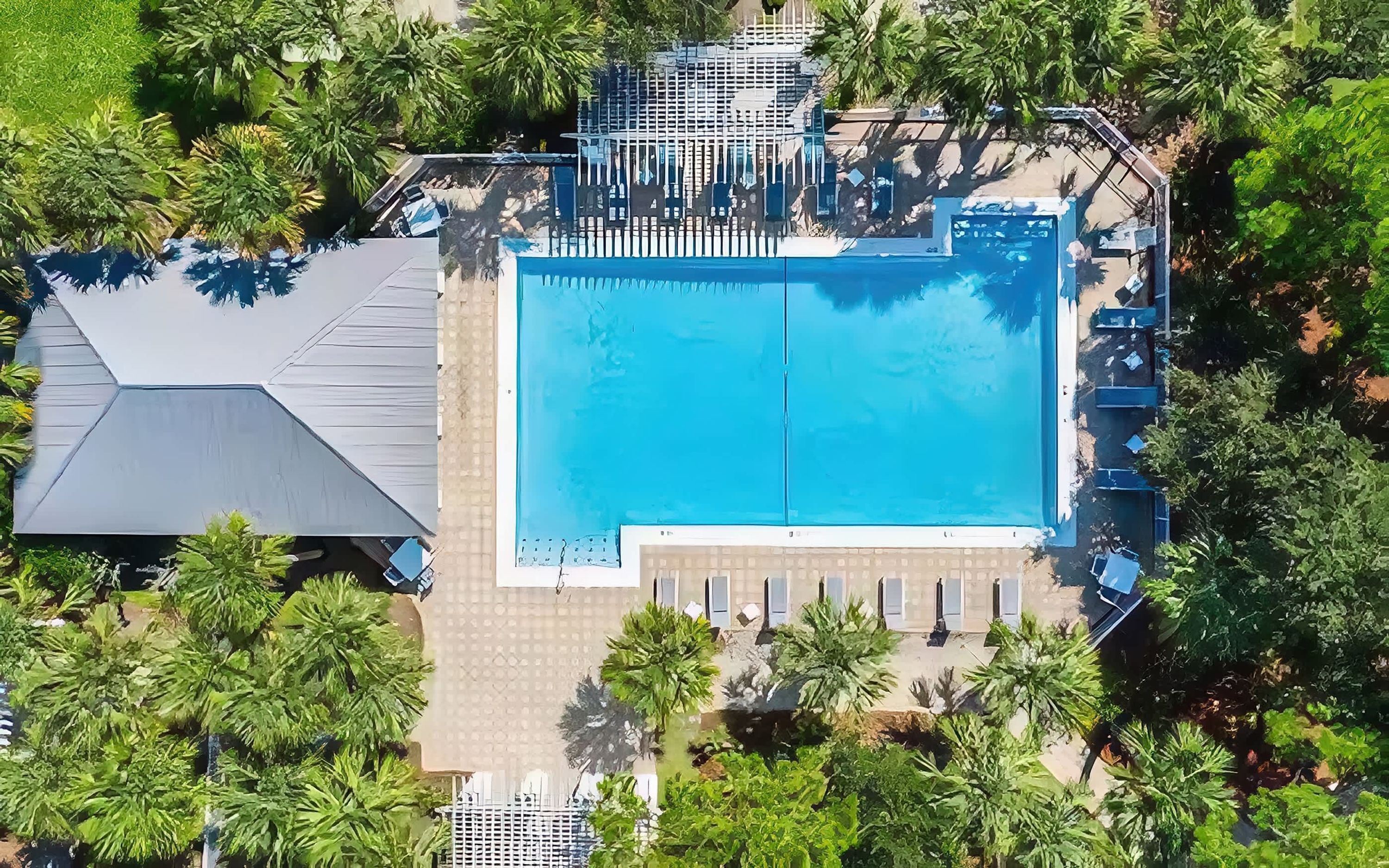 Vue aérienne d’un complexe hôtelier doté d’une piscine centrale, entourée de palmiers et d’un espace abrité avec des chaises longues.