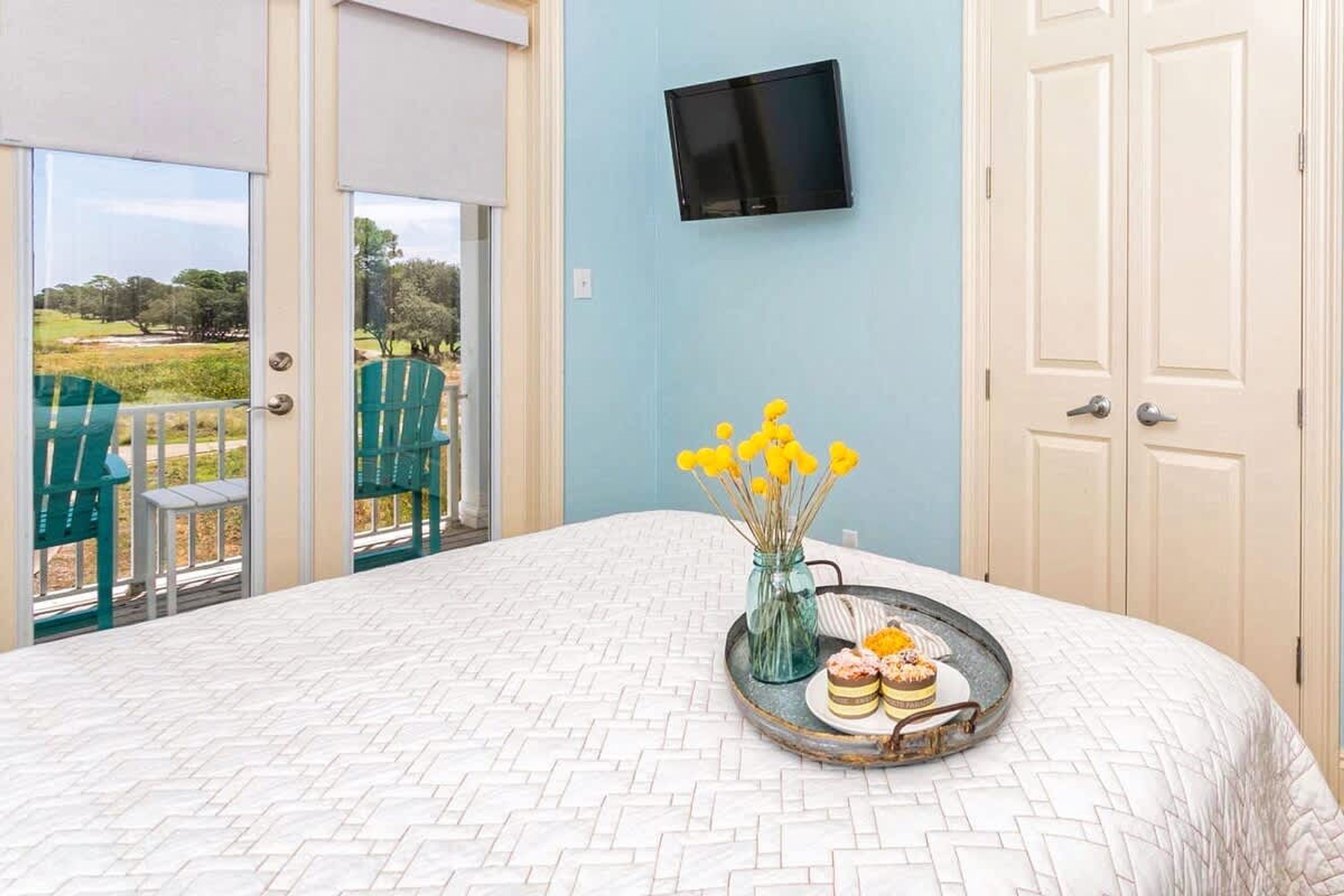 Ein Schlafzimmer mit einem Bett, einem Tablett mit Desserts, einer Vase mit gelben Blumen und einem Fernseher an der Wand.