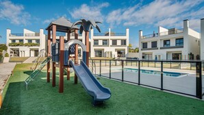 Un terrain de jeux équipé d’un toboggan et de structures d’escalade, entouré de bâtiments modernes et d’une piscine.